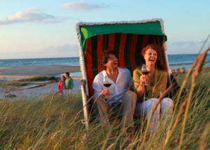 Paar genießt den Abend im Strandkorb am Meer. Paar im Strandkorb an der Ostsee, lachend mit Weingläsern in der Hand bei Sonnenuntergang.