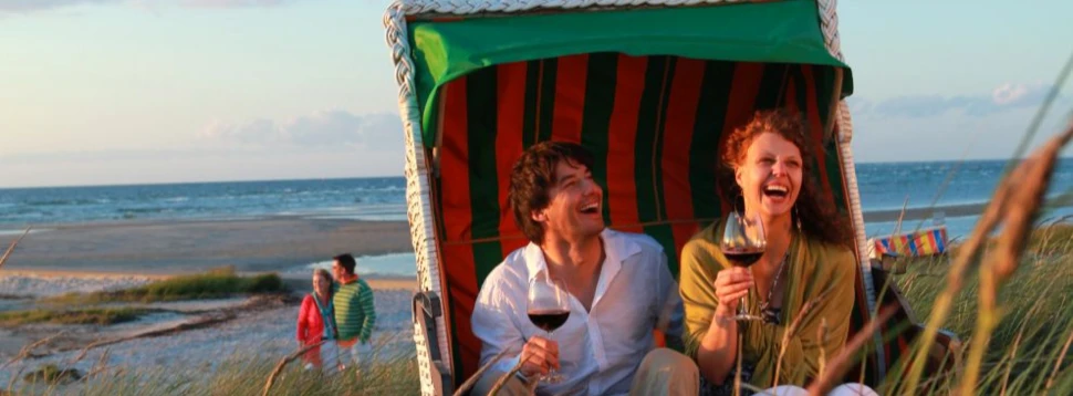 Paar im Strandkorb an der Ostsee, lachend mit Weingläsern in der Hand bei Sonnenuntergang.