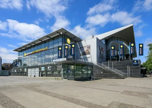 Foto von einem großen Gebäude, wo von Außen Flaggen und Aufschriften mit dem Namen "Merkur Ostseehallen" zu sehen sind