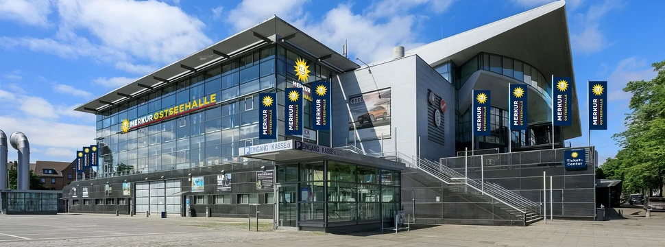 Foto von einem großen Gebäude, wo von Außen Flaggen und Aufschriften mit dem Namen "Merkur Ostseehallen" zu sehen sind
