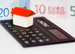 Ein kleines Haus steht auf einem Taschenrechner, im Hintergrund liegt Geld