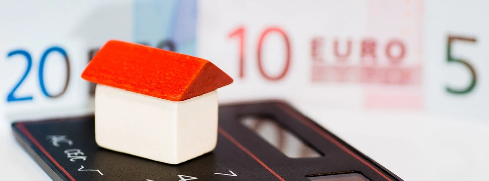 Ein kleines Haus steht auf einem Taschenrechner, im Hintergrund liegt Geld