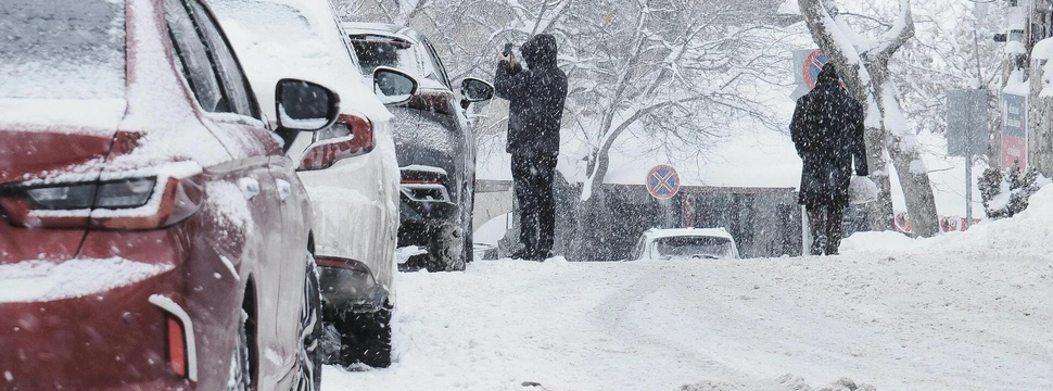 Mehrere zugeschneite Autos auf einer Straße, die von Menschen vom Eis befreit werden