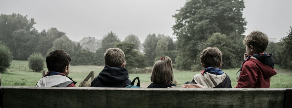 Fünf Kinder sitzen nebeneinander auf einer Bank. Das Foto ist von hinten aufgenommen.
