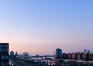 Blick auf einen Hafen bei Dämmerung mit moderner Architektur, Wasser und einem leicht rosafarbenen Himmel.