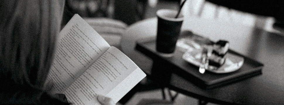 Frau sitzt mit einem Buch vor einem kleinen Tisch, auf dem ein Becher und ein Stück Kuchen stehen