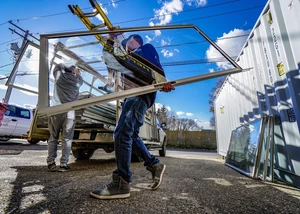 Handwerker bei der Glasmontage auf einem Gewerbehof unter blauem Himmel. Zwei Handwerker in Arbeitskleidung montieren gemeinsam große Glasscheiben bei blauem Himmel auf einem Bauhof.