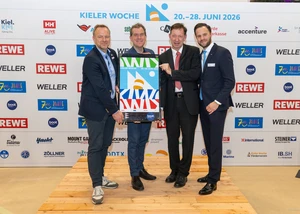 Vier Männer stehen vor einer Kieler Woche Werbewand und halten das neue Kieler Woche Plakat in den Händen