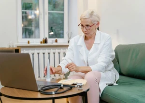 Eine Frau mit grauen Haaren auf einem grünen Sofa vor einem Laptop, das auf einem Tisch steht