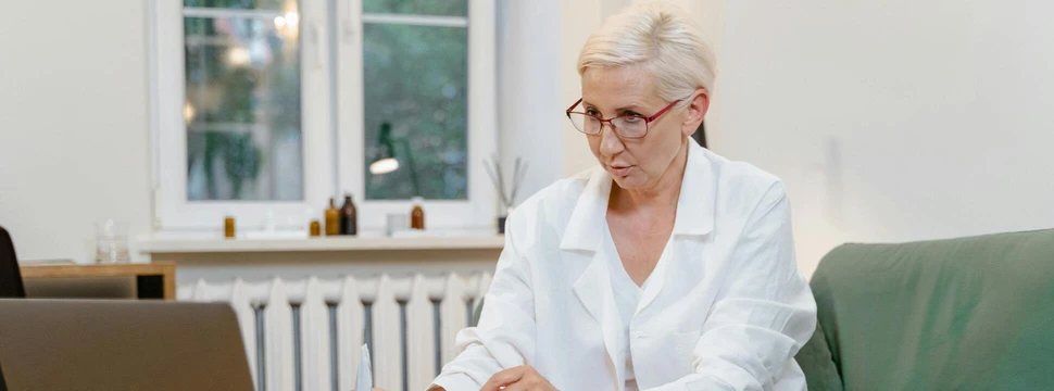 Eine Frau mit grauen Haaren auf einem grünen Sofa vor einem Laptop, das auf einem Tisch steht