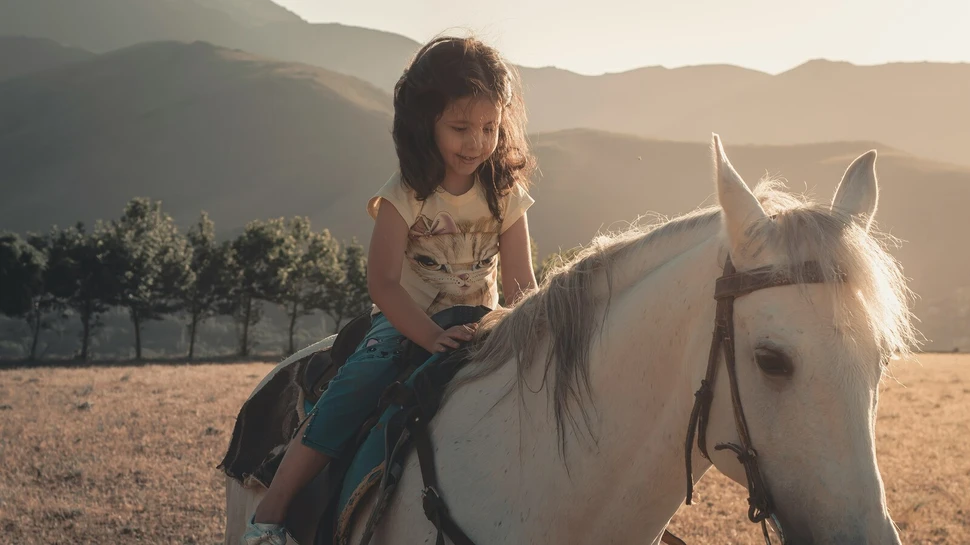 kleines Mädchen sitzt auf einem weißen Pferd. Im Hintergrund sieht man eine Berglandschaften