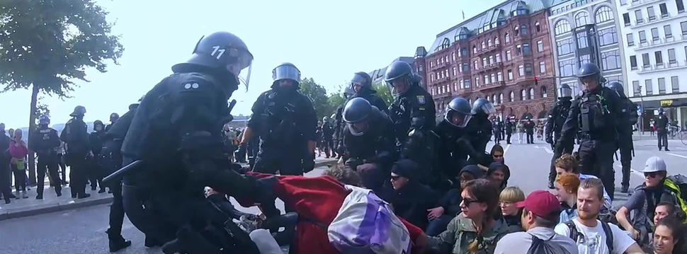 Polizisten ziehen eine Person aus einer Menschengruppe, die auf den Boden sitzen