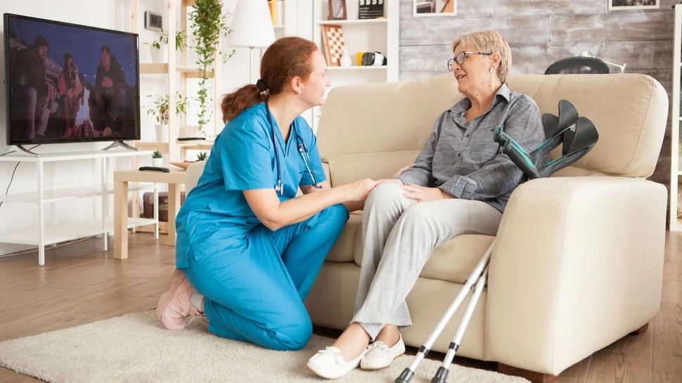 Vertrauensvolle Pflege im eigenen Zuhause. Pflegerin spricht lächelnd mit älterer Frau auf Sofa in hellen Wohnzimmer.